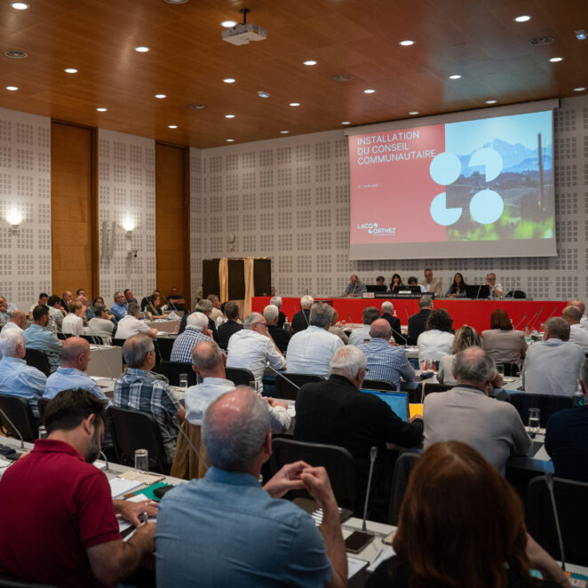 Salle de réunion remplie avec face à nous les élus, de dos, assis en rangées, se tenant face au bureau principal et à un diaporama qui défile grâce à un projecteur.