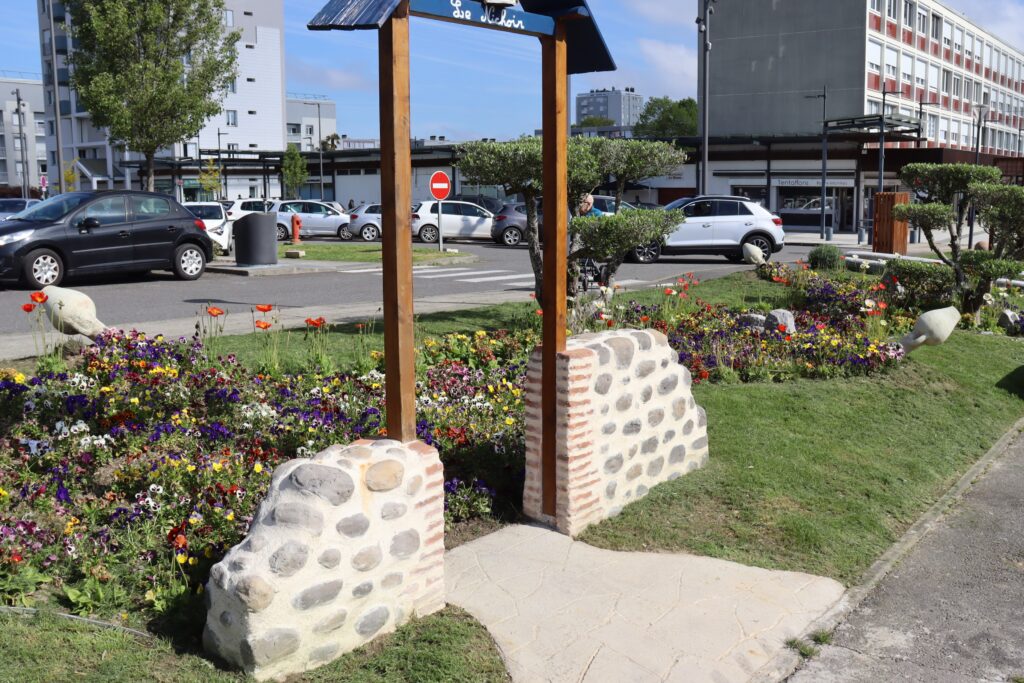 Face à nous se dresse un petit espace vert décoré d'un nichoir en hauteur et de plein de fleurs colorées de gauche à droite de la photo, on distingue aussi des arbustes bien taillés, et en fond des voitures garées et des immeubles, montrant que nous somme en pleine ville.