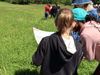 Enfant de dos avec un pull à capuche qui regarde sa feuille d'école avec une autre élève avec une casquette à sa droite, les filles sont dans un espace naturel, et plus loin devant elle on distingue d'autres groupes d'enfants regardant leur feuille de travail également.