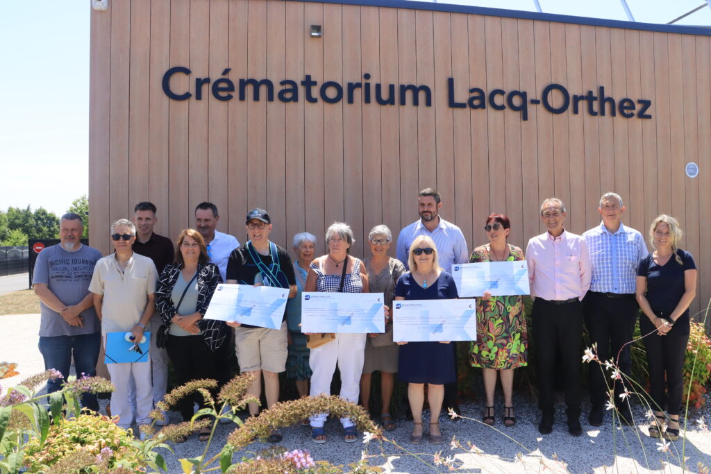 Groupe d'une dizaine de personnes alignées devant la façade où il est écrit dessus "Crématorium Lacq-Orthez" avec en premier plan, une personne représentant chaque association et tenant dans sa main un chèque géant.